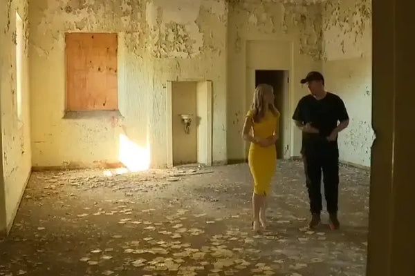 Brett Patman and Samantha Brett walk through a decaying room inside Callan Park, surrounded by peeling paint and crumbling walls, during a 7News Sydney feature on Lost Collective.