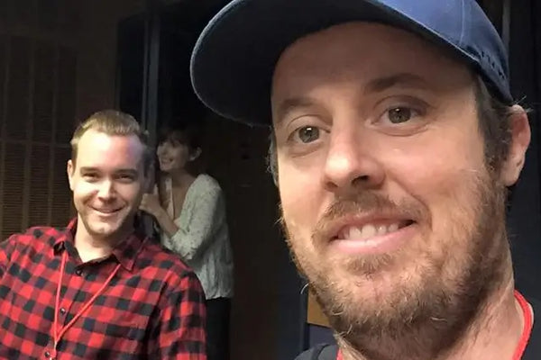 Brett Patman in a navy cap in the foreground, with Tim Frawley in a red and black flannel shirt behind him giving a thumbs up in the ABC Radio National studio. A third person partially visible in the doorway.
