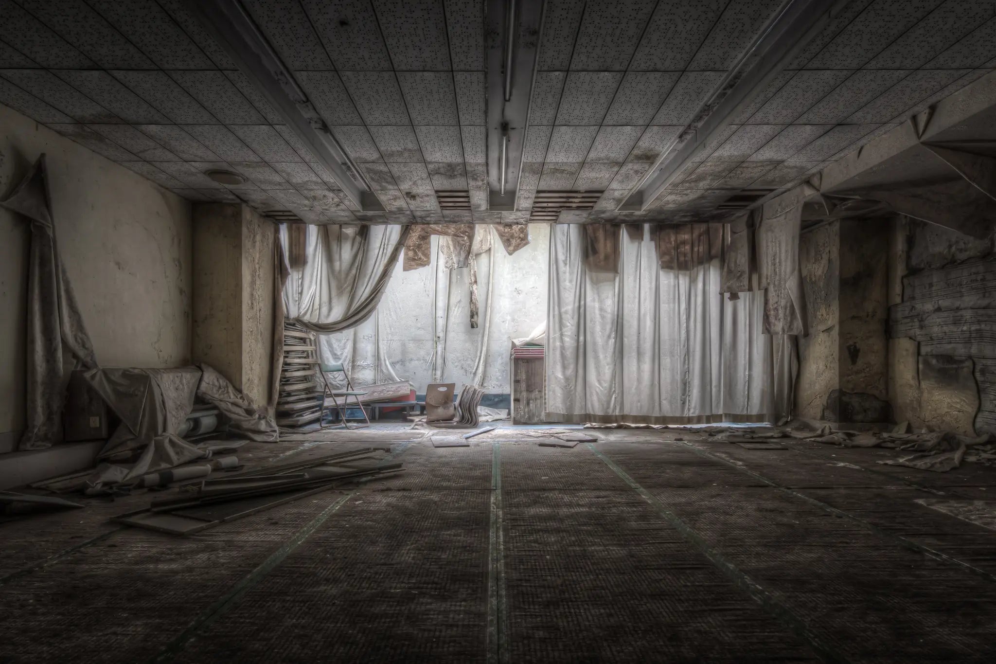 An empty function room inside Kinugawa Kan, cream fabric drapes fallen from the ceiling, a step ladder against the back wall, debris scattered across the grid-tiled floor.