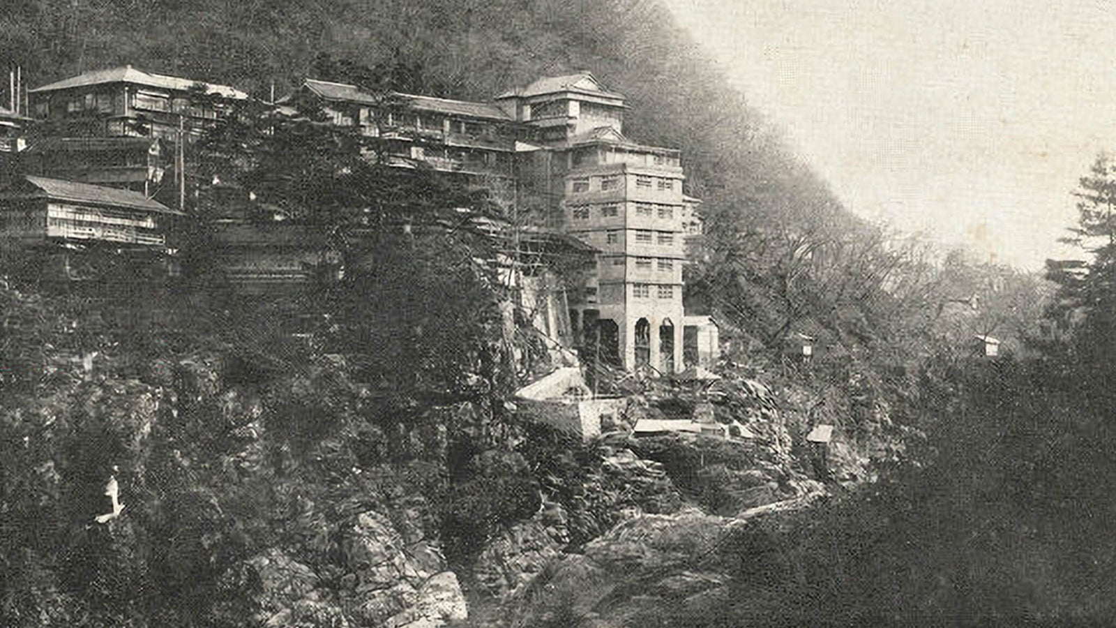 Historical black and white photograph of Kinugawa Kan on the cliffs above the Kinugawa River gorge, the multi-storey hotel and its traditional Japanese annexes terraced down the rocky slope.