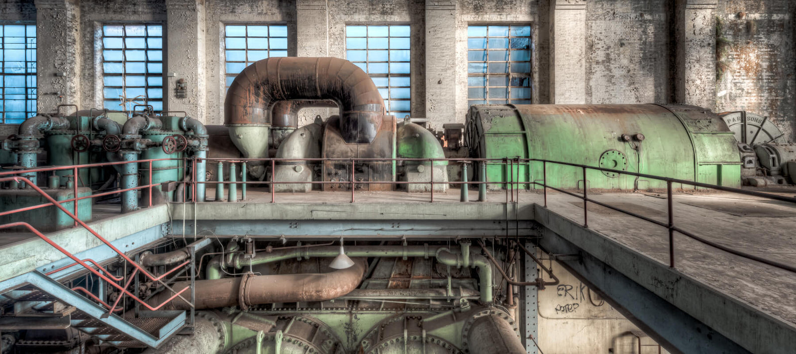 An industrial interior featuring large rusted machinery, green turbines, and a brick wall backdrop with tall windows, showcasing the grandeur of abandoned industry.