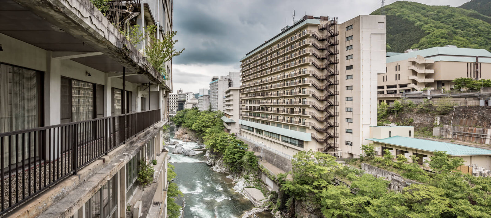 View of a river gorge flanked by an abandoned hotel with weathered balconies and a modern building, surrounded by lush greenery and distant mountains in Japan.