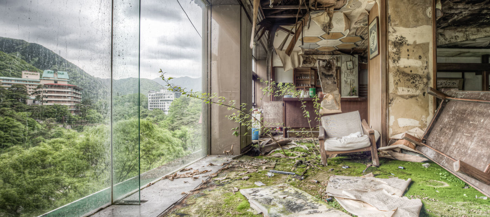 A decayed room in Kinugawa Kan, an abandoned hotel in Kinugawa Onsen, Japan. Moss covers the floors, vegetation grows inside, and large glass windows reveal green mountains and derelict buildings along the Kinugawa River.