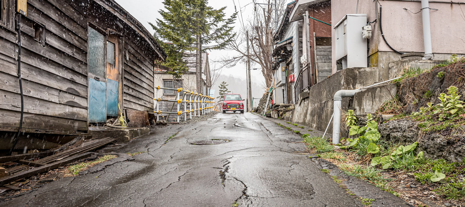 A quiet, narrow street in Yubari, Japan, featuring weathered wooden buildings, a red car parked at the end, and light snow falling.