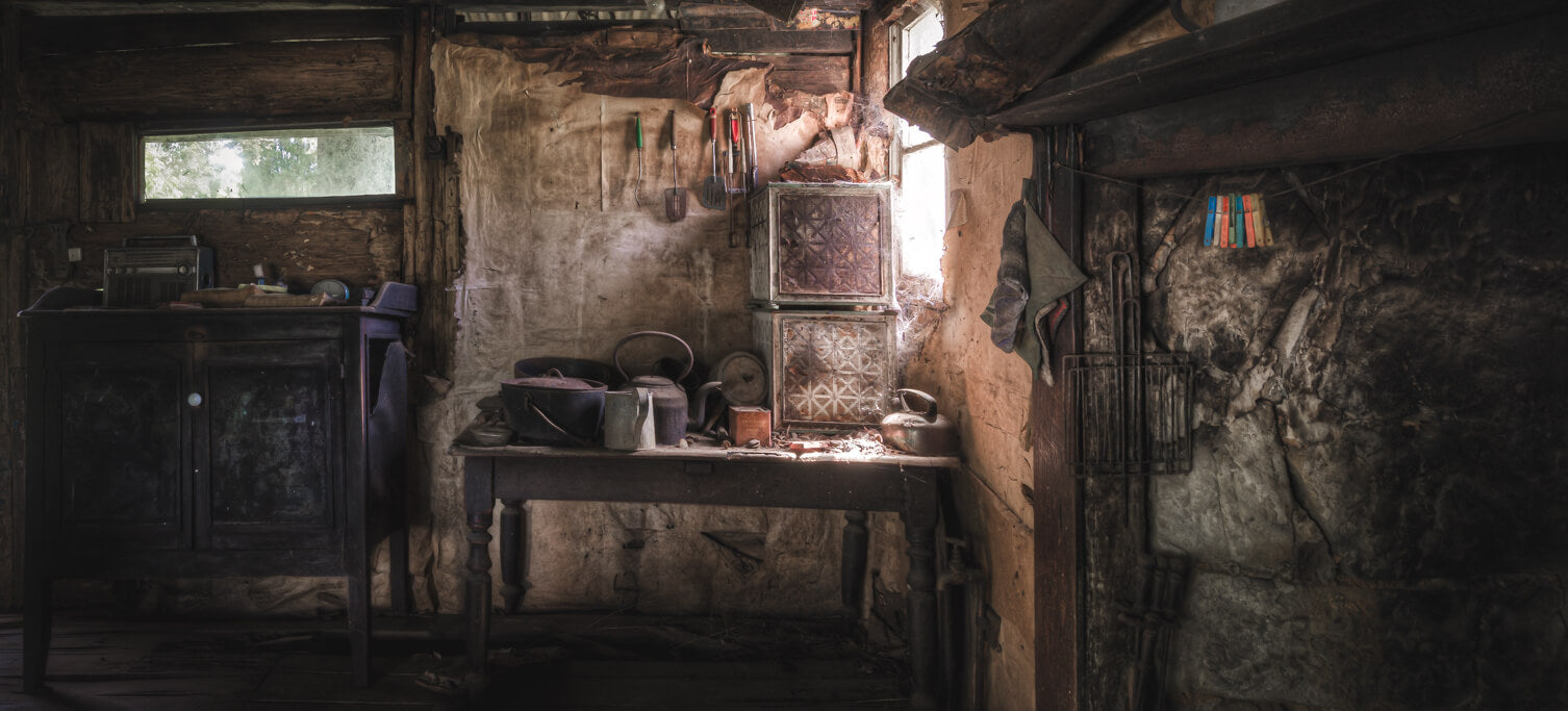 Interior of an abandoned rural homestead showcasing vintage kitchen tools, an aged cupboard, and rustic decor bathed in natural light through a small window.