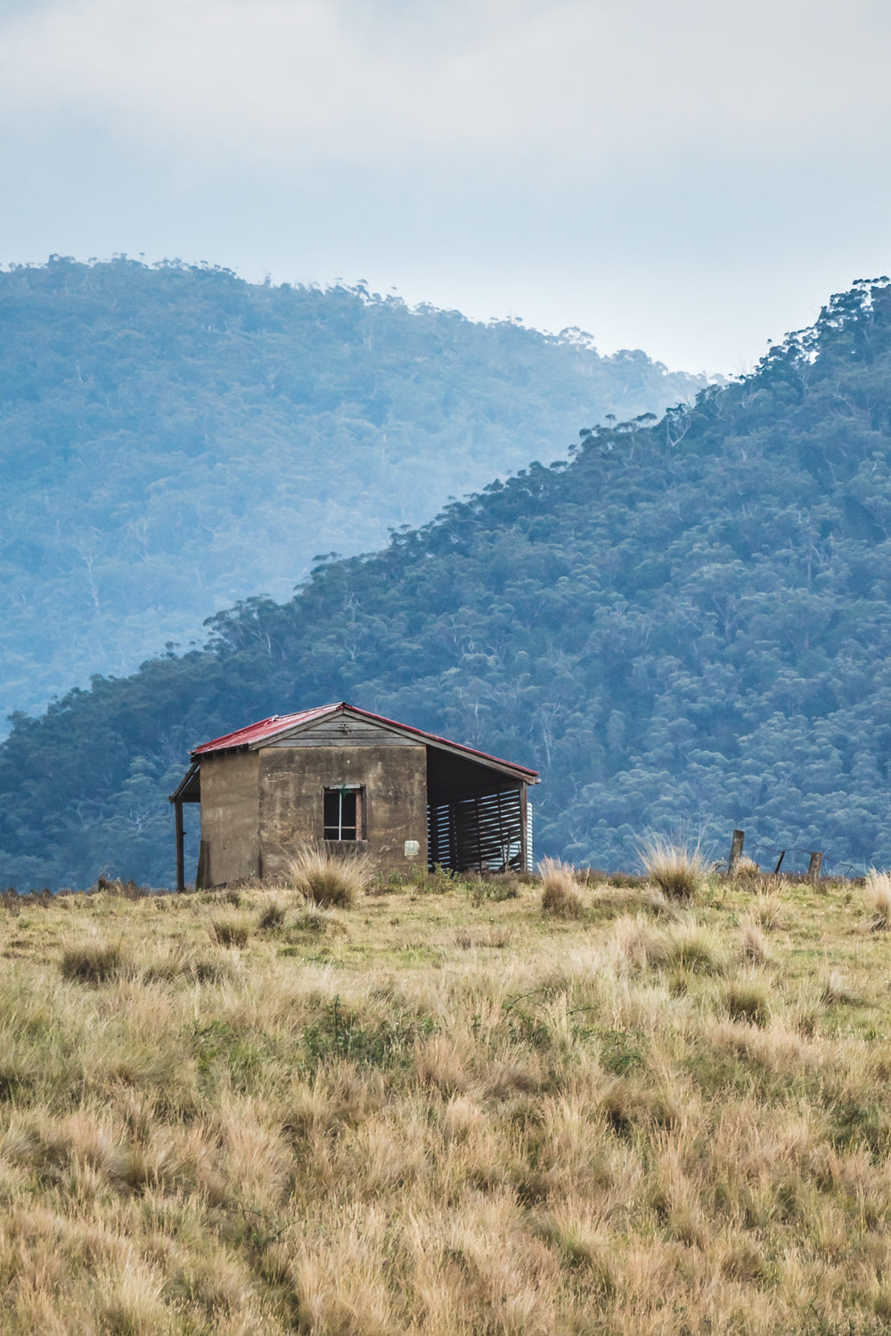 A solitary weathered house with a red roof perched on a hill, surrounded by high country grasslands and mountains in the background.