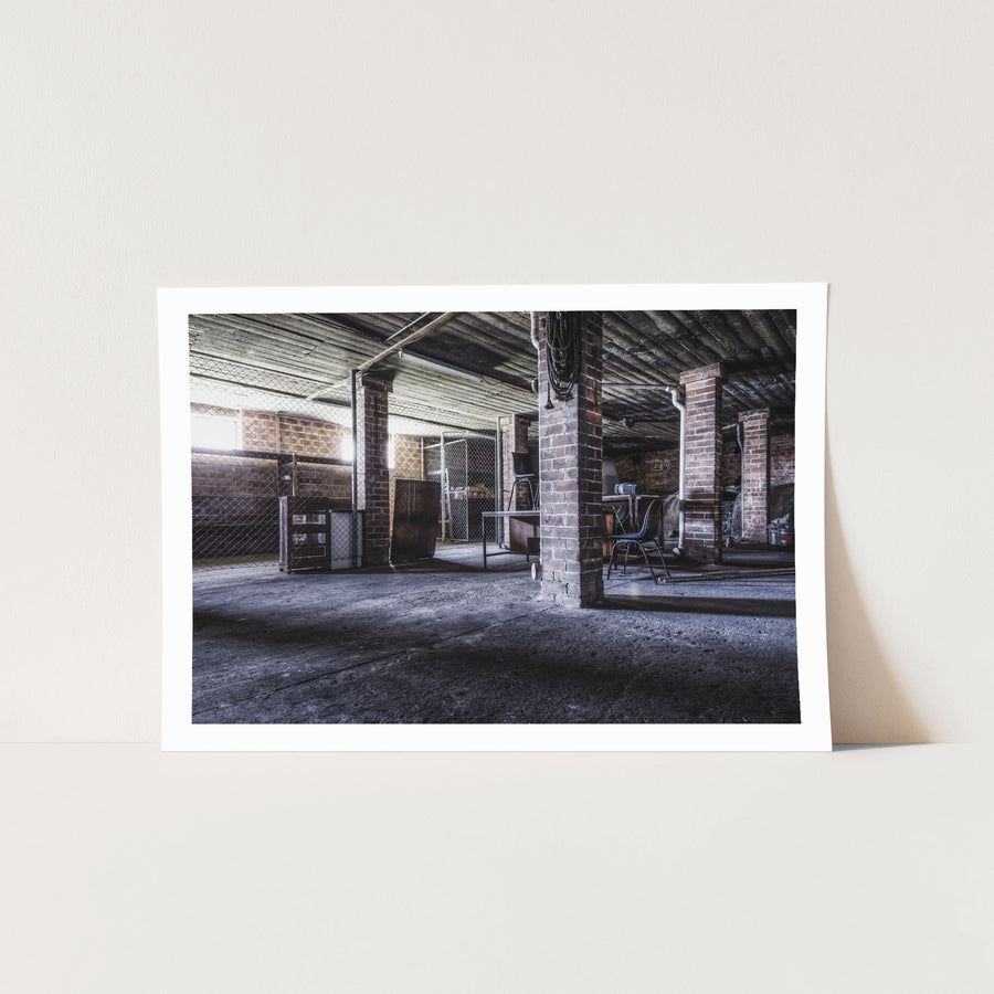A coiled rope hangs from a brick column in the basement of the administration building at Awaba Colliery, with an overturned office chair on a desk and shadows across the concrete floor.