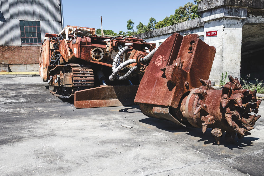 Rust-covered rotary cutting head of a boring machine on a concrete slab at Awaba Colliery, with white hydraulic hoses still connected and a corrugated iron building visible behind.