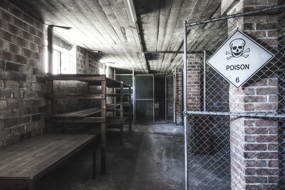 A Poison 6 diamond hazard sign on a chain-link gate marks the entrance to the chemical storage corridor at Awaba Colliery, with brick walls, timber ceiling, and empty wooden shelving visible beyond.