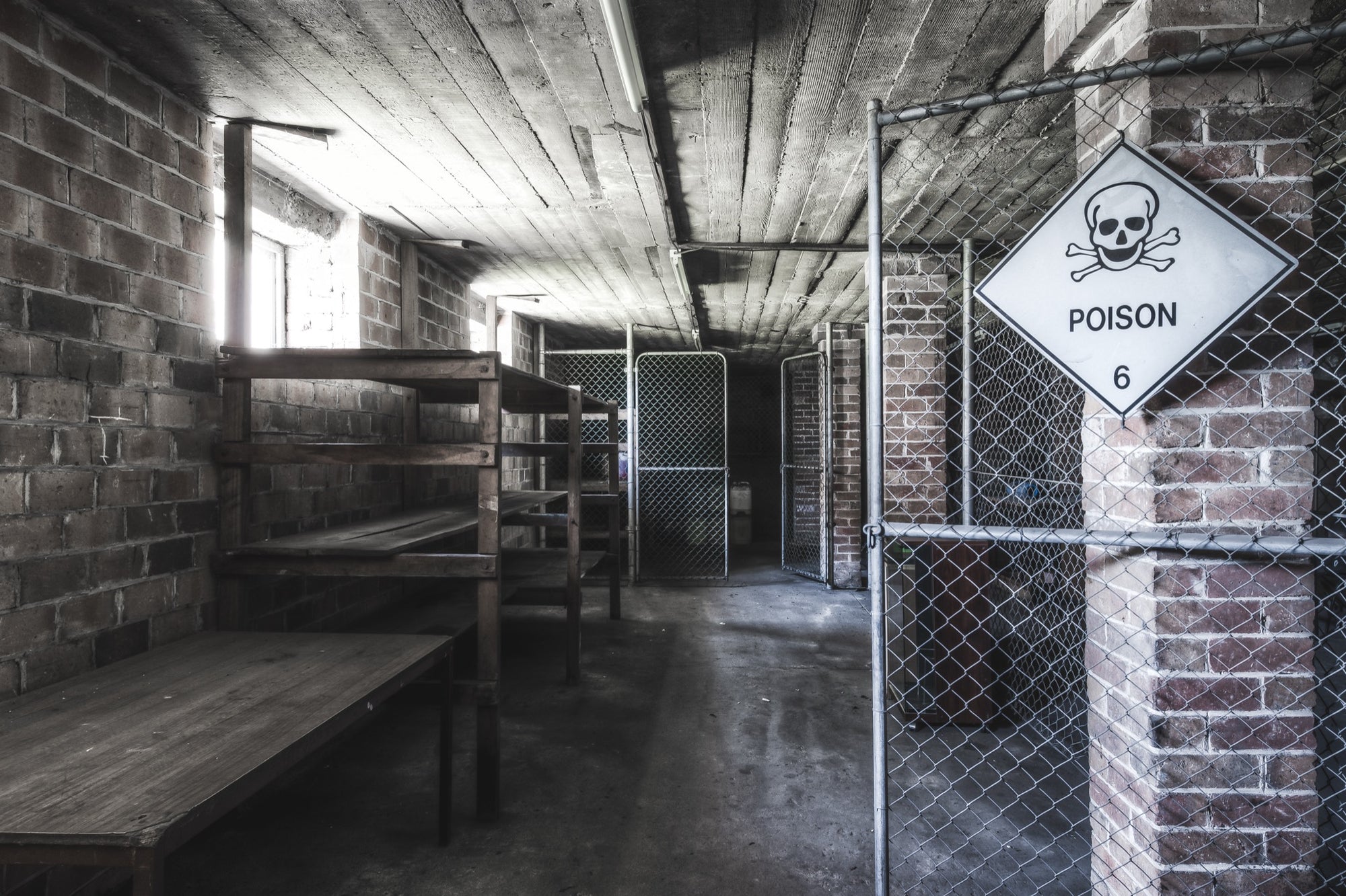 A Poison 6 diamond hazard sign on a chain-link gate marks the entrance to the chemical storage corridor at Awaba Colliery, with brick walls, timber ceiling, and empty wooden shelving visible beyond.