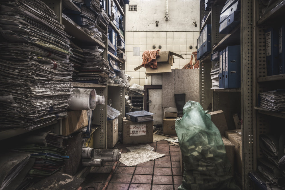 Cardboard boxes and paper stacked on metal shelving in a former shower room at Awaba Colliery, with the original shower heads still visible on the cracked tiled back wall.