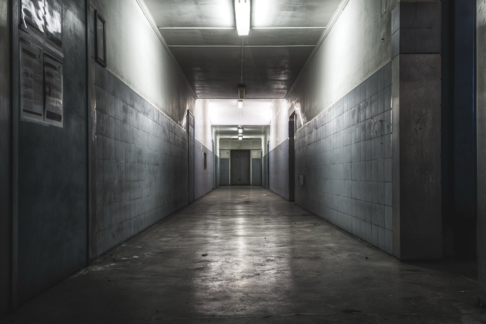 A narrow corridor at Awaba Colliery with fluorescent lights still burning along the ceiling, light-grey tiled walls, a concrete floor, and a closed door at the far end.