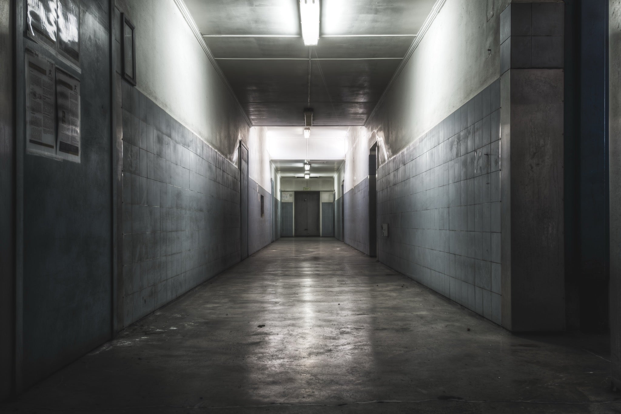 A narrow corridor at Awaba Colliery with fluorescent lights still burning along the ceiling, light-grey tiled walls, a concrete floor, and a closed door at the far end.