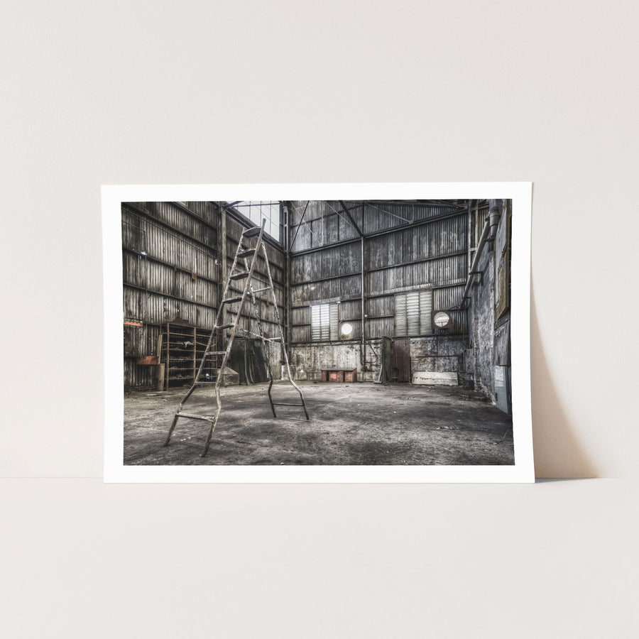 A rusted A-frame ladder stands in a workshop interior at Awaba Colliery, with a wooden shelving unit, a grinder sign, and a high louvred window letting in the only natural light.