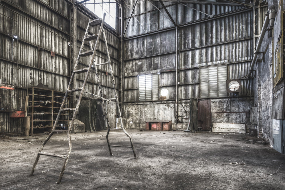 A rusted A-frame ladder stands in a workshop interior at Awaba Colliery, with a wooden shelving unit, a grinder sign, and a high louvred window letting in the only natural light.