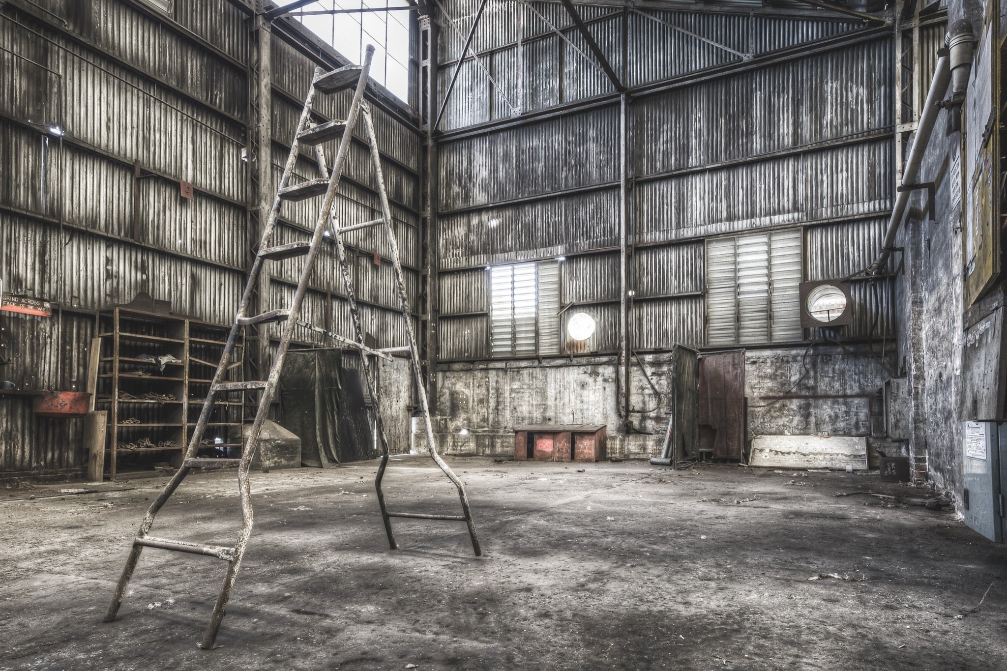 A rusted A-frame ladder stands in a workshop interior at Awaba Colliery, with a wooden shelving unit, a grinder sign, and a high louvred window letting in the only natural light.