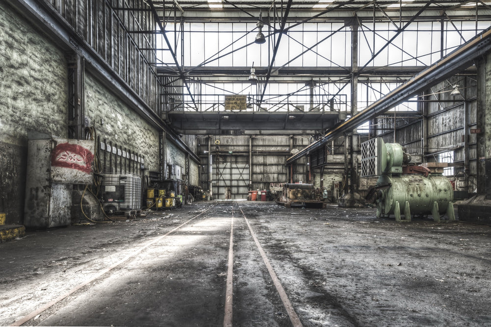 Rusted cylindrical machinery and twin rail tracks on the workshop floor at Awaba Colliery, with a Johnson-Hepburn sign hanging from a gantry crane and grease buckets lining the left wall.