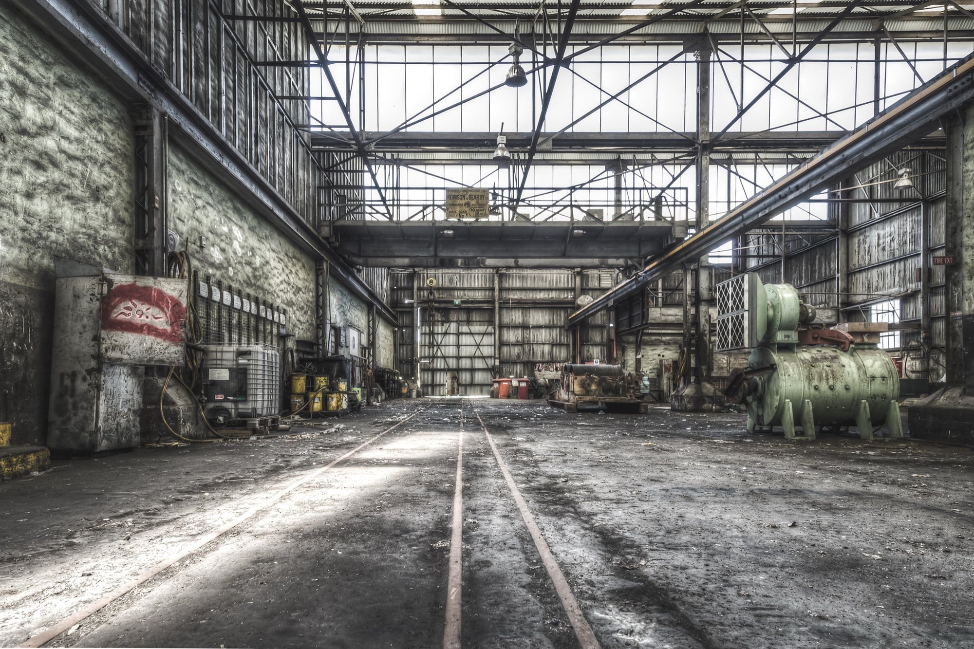 Rusted cylindrical machinery and twin rail tracks on the workshop floor at Awaba Colliery, with a Johnson-Hepburn sign hanging from a gantry crane and grease buckets lining the left wall.