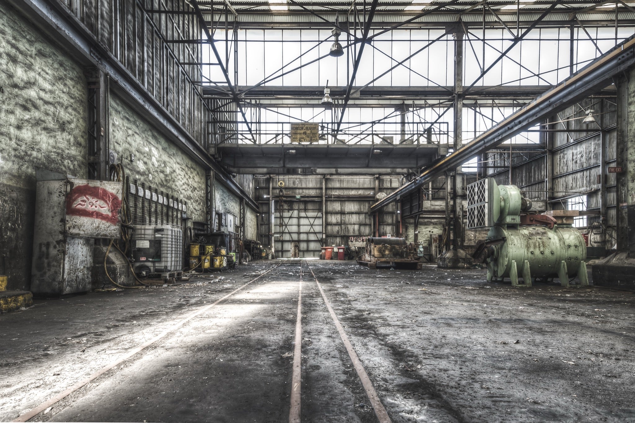 Rusted cylindrical machinery and twin rail tracks on the workshop floor at Awaba Colliery, with a Johnson-Hepburn sign hanging from a gantry crane and grease buckets lining the left wall.