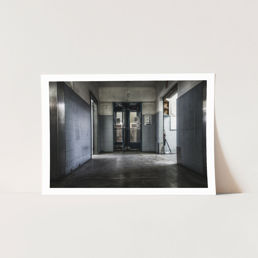 A tiled hallway at Awaba leads to a pair of partially open security-grated double doors, with a text sign on the right wall and a mop and bucket visible through the gap.
