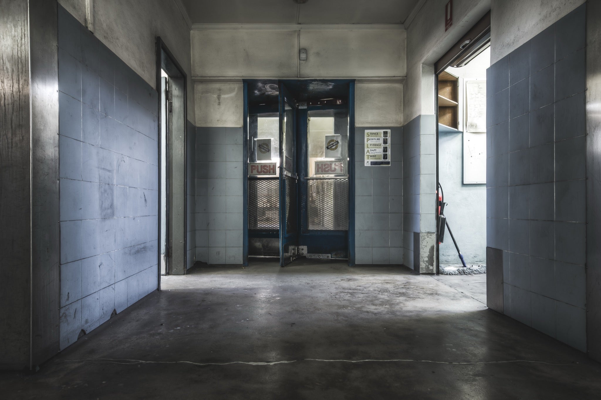 A tiled hallway at Awaba leads to a pair of partially open security-grated double doors, with a text sign on the right wall and a mop and bucket visible through the gap.