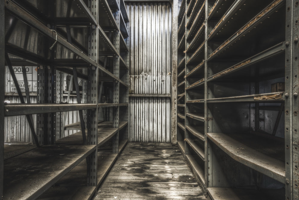 Rust-streaked metal shelving lining a narrow aisle in a derelict storeroom at Awaba Colliery, with warped floorboards and a partially legible sign visible on the left unit.