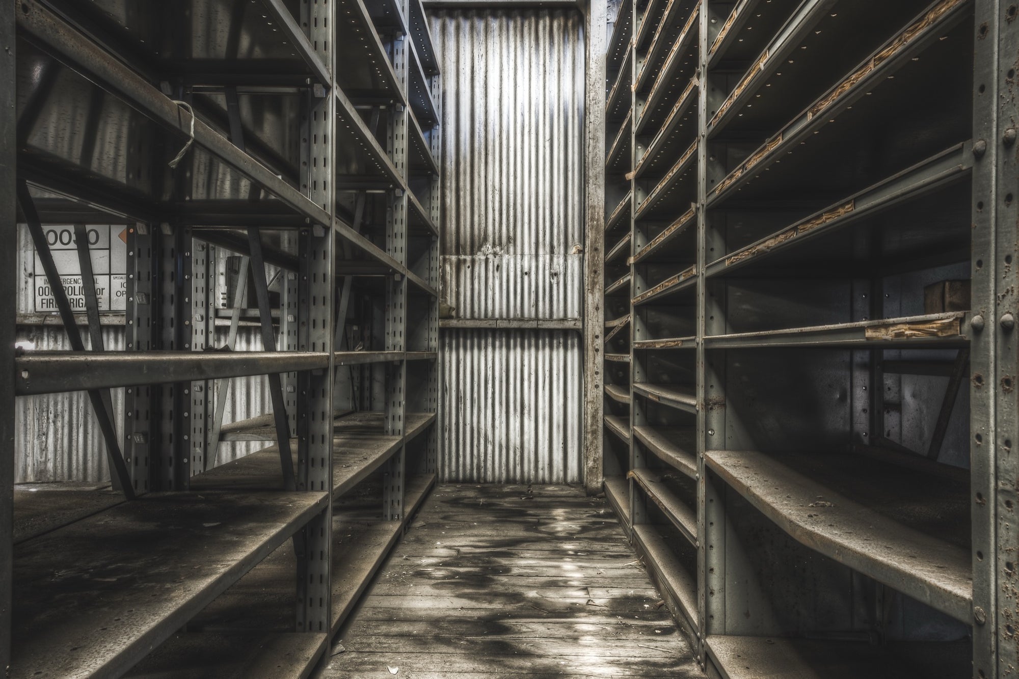 Rust-streaked metal shelving lining a narrow aisle in a derelict storeroom at Awaba Colliery, with warped floorboards and a partially legible sign visible on the left unit.