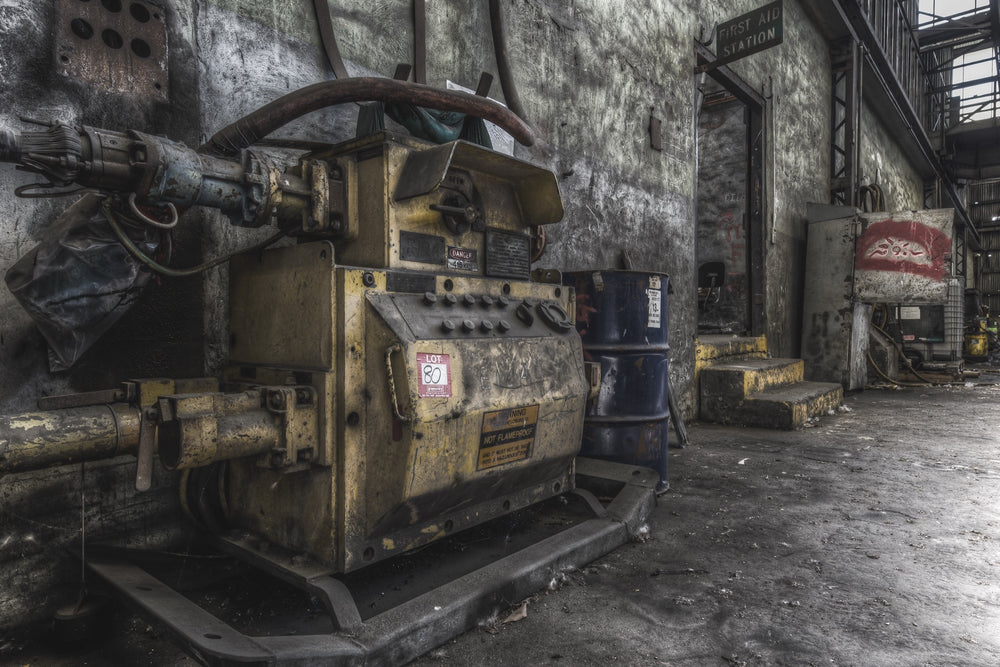 A yellow slurry pump tagged 'Lot 80' with control knobs and pipework in the interior of Awaba Colliery, blue barrels stacked against the wall and a First Aid Station doorway visible in the background.