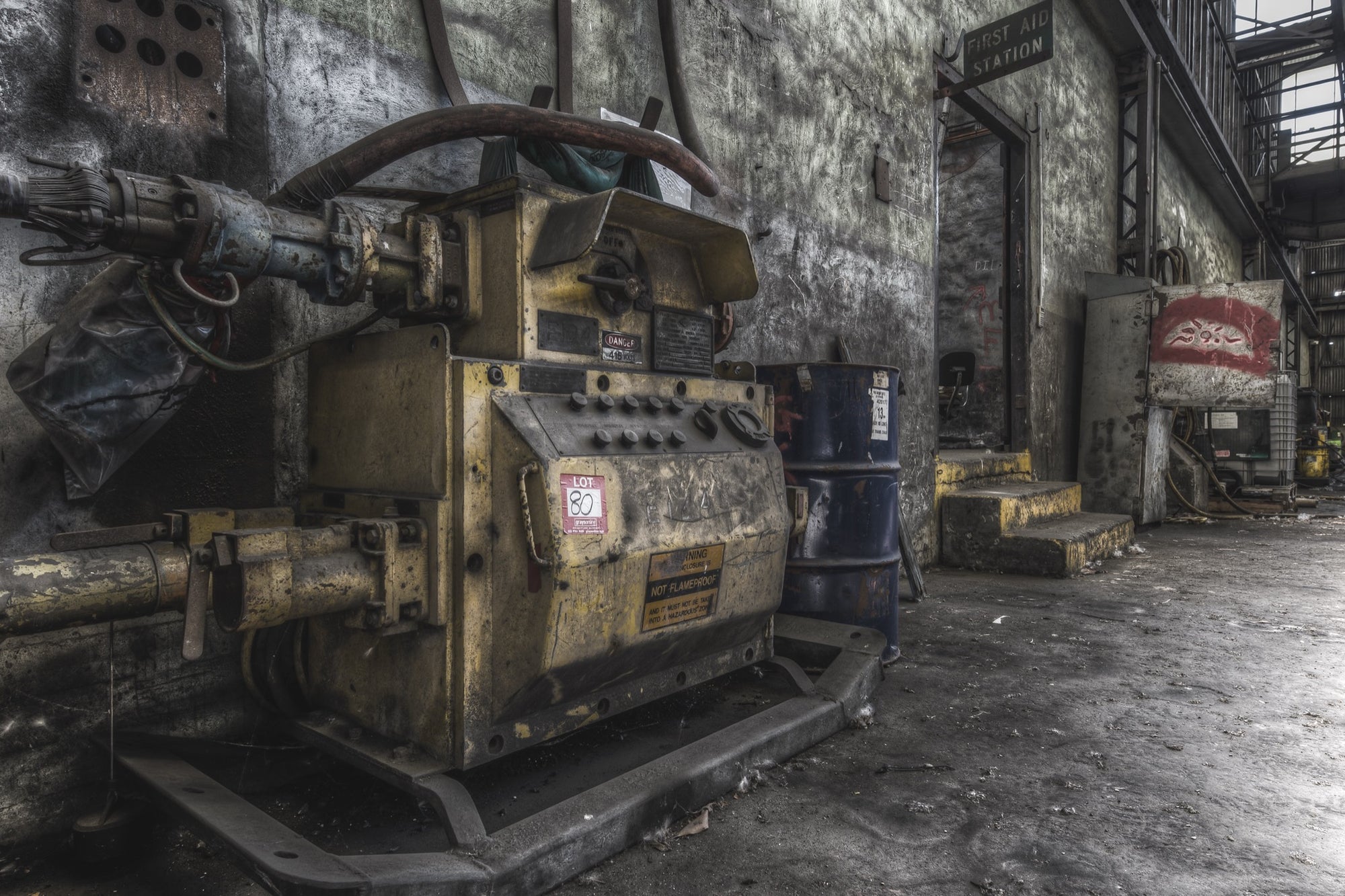 A yellow slurry pump tagged 'Lot 80' with control knobs and pipework in the interior of Awaba Colliery, blue barrels stacked against the wall and a First Aid Station doorway visible in the background.