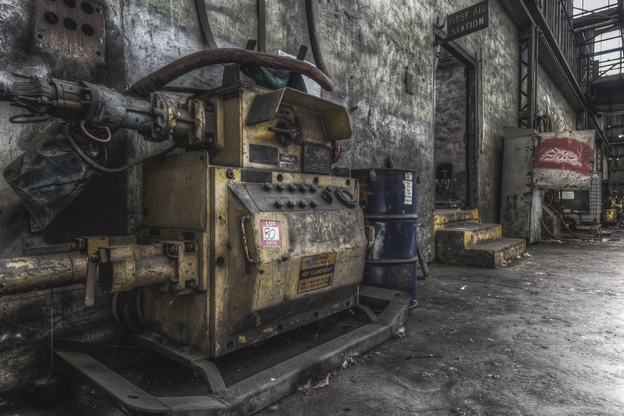 A yellow slurry pump tagged 'Lot 80' with control knobs and pipework in the interior of Awaba Colliery, blue barrels stacked against the wall and a First Aid Station doorway visible in the background.