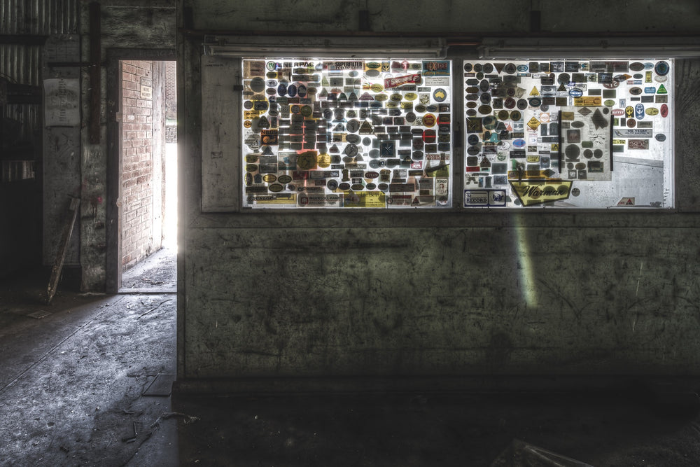 Sticker-covered storeman's office windows at Awaba Colliery, with brand labels for Ingersoll Rand, Exide, and Superfilter legible against backlit glass and a rectangle of light on the wall below.
