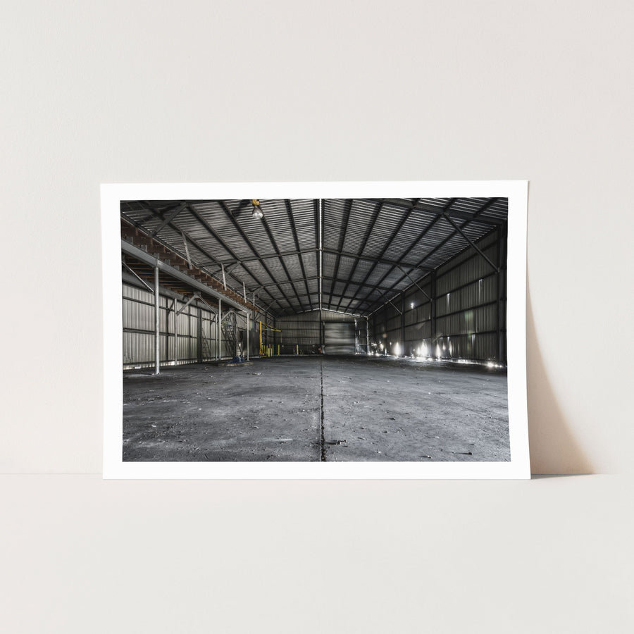 Interior of the storage shed at Awaba Colliery, showing buckled corrugated metal walls, a metal walkway with timber steps along the left side, a yellow gantry in the mid-ground, and bright window light cutting across a concrete floor.