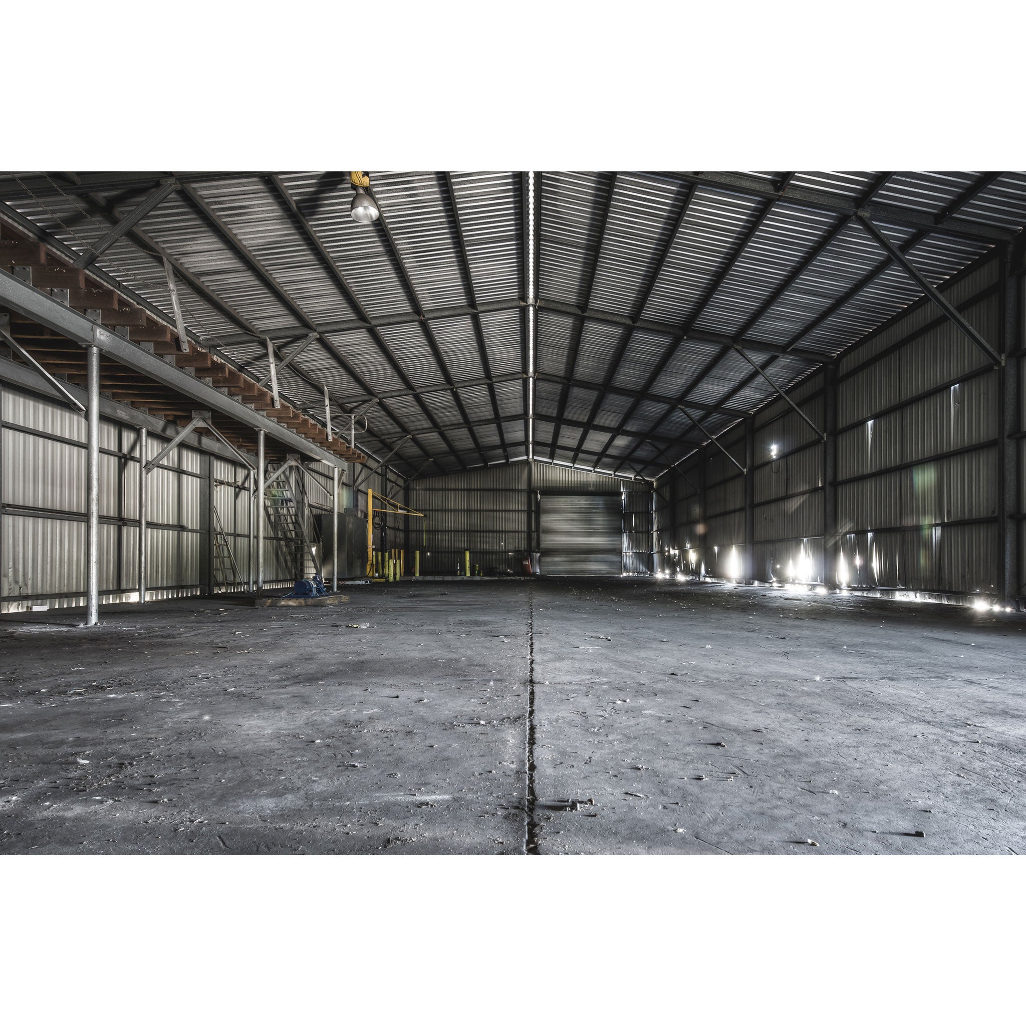 Interior of the storage shed at Awaba Colliery, showing buckled corrugated metal walls, a metal walkway with timber steps along the left side, a yellow gantry in the mid-ground, and bright window light cutting across a concrete floor.