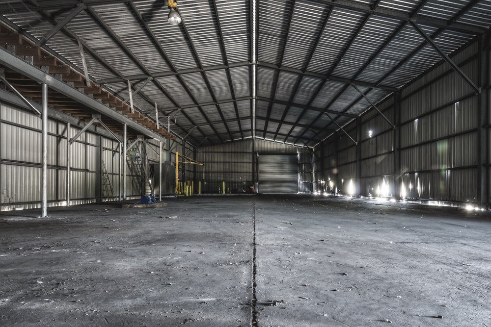 Interior of the storage shed at Awaba Colliery, showing buckled corrugated metal walls, a metal walkway with timber steps along the left side, a yellow gantry in the mid-ground, and bright window light cutting across a concrete floor.