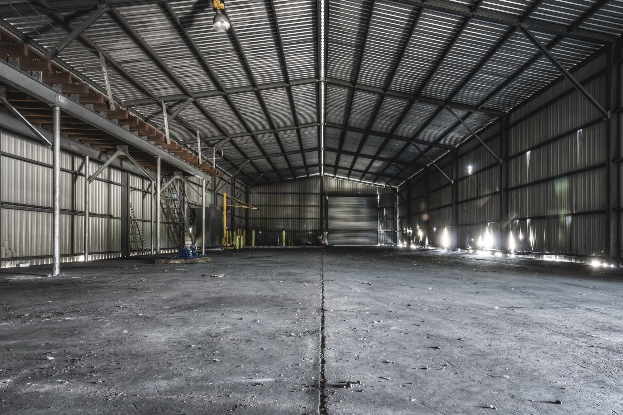 Interior of the storage shed at Awaba Colliery, showing buckled corrugated metal walls, a metal walkway with timber steps along the left side, a yellow gantry in the mid-ground, and bright window light cutting across a concrete floor.