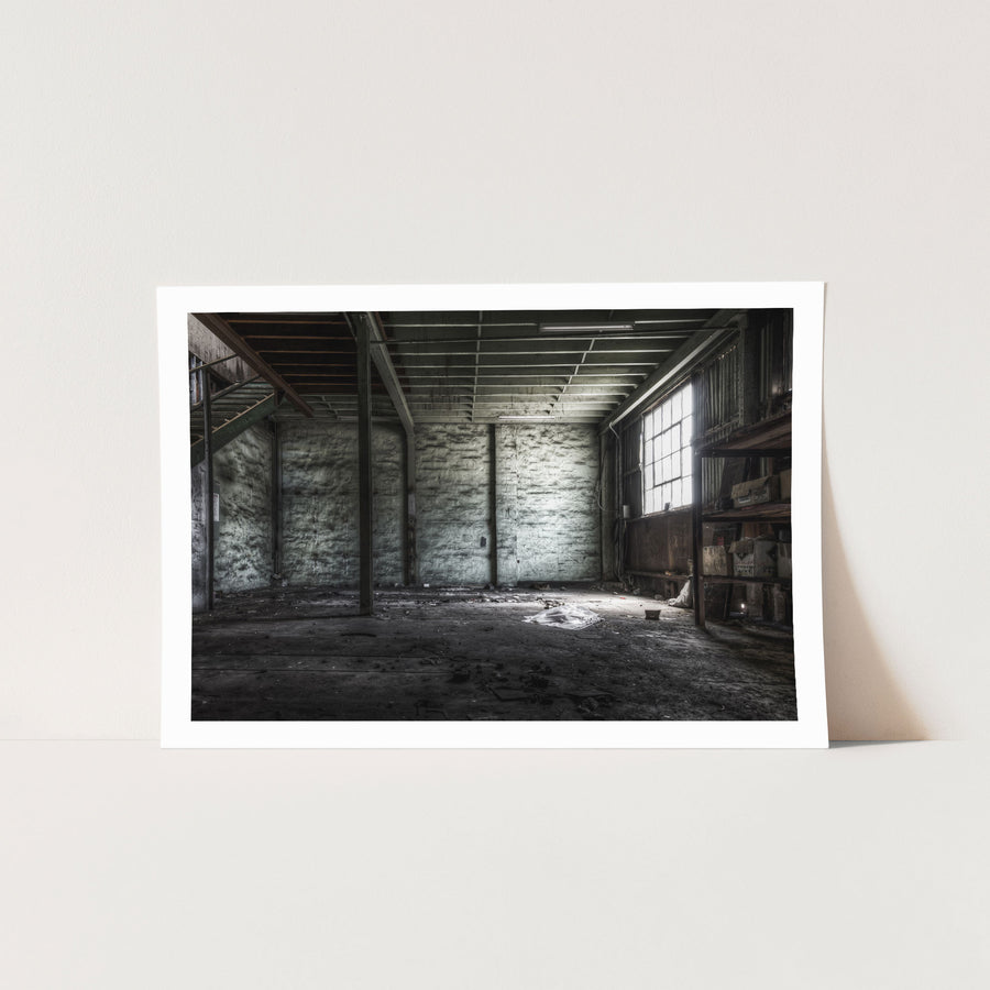 Cardboard boxes on timber shelving beside a grimy multi-pane window in the main store at Awaba Colliery, with white plastic sheeting on the concrete floor and a metal staircase rising at left.