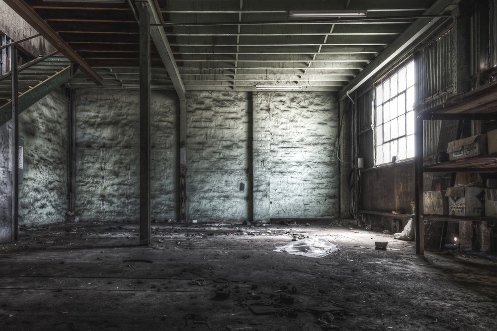 Cardboard boxes on timber shelving beside a grimy multi-pane window in the main store at Awaba Colliery, with white plastic sheeting on the concrete floor and a metal staircase rising at left.