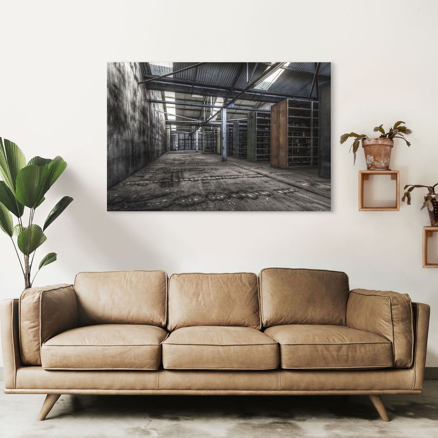 Emptied metal shelving units receding through the upper storeroom at Awaba Colliery, with skylights in a corrugated iron roof casting light across water-stained concrete walls and worn timber floorboards.