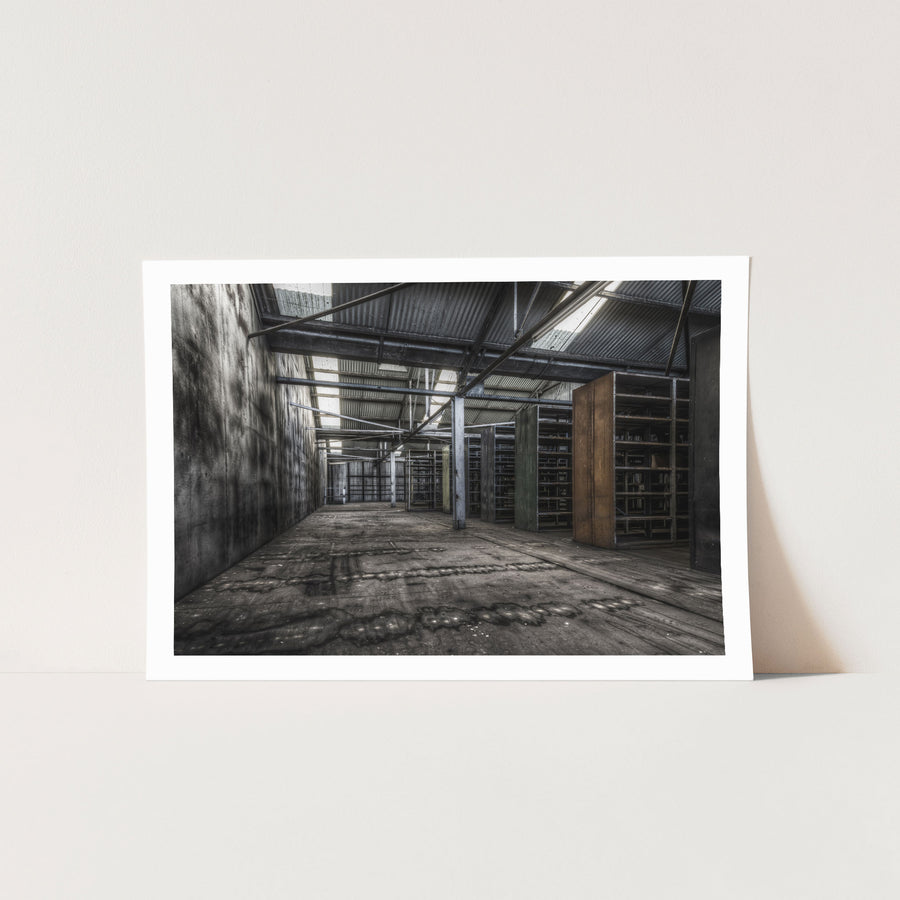 Emptied metal shelving units receding through the upper storeroom at Awaba Colliery, with skylights in a corrugated iron roof casting light across water-stained concrete walls and worn timber floorboards.