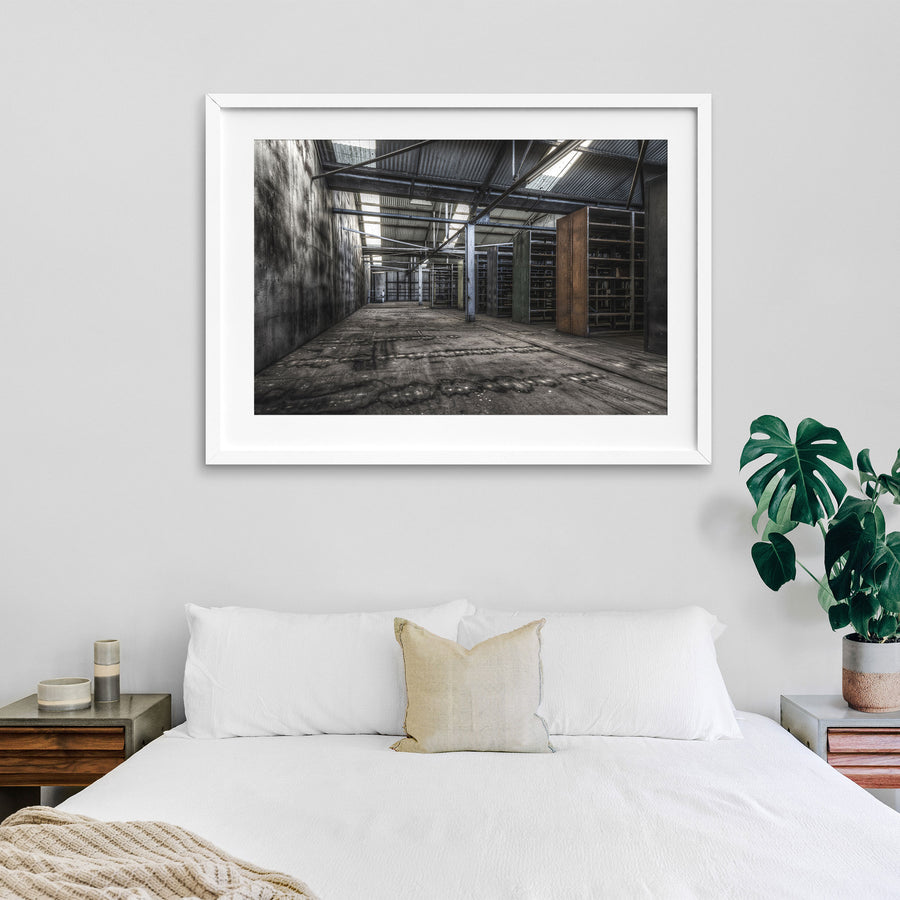 Emptied metal shelving units receding through the upper storeroom at Awaba Colliery, with skylights in a corrugated iron roof casting light across water-stained concrete walls and worn timber floorboards.