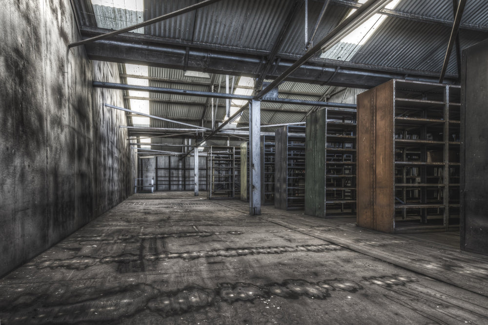 Emptied metal shelving units receding through the upper storeroom at Awaba Colliery, with skylights in a corrugated iron roof casting light across water-stained concrete walls and worn timber floorboards.