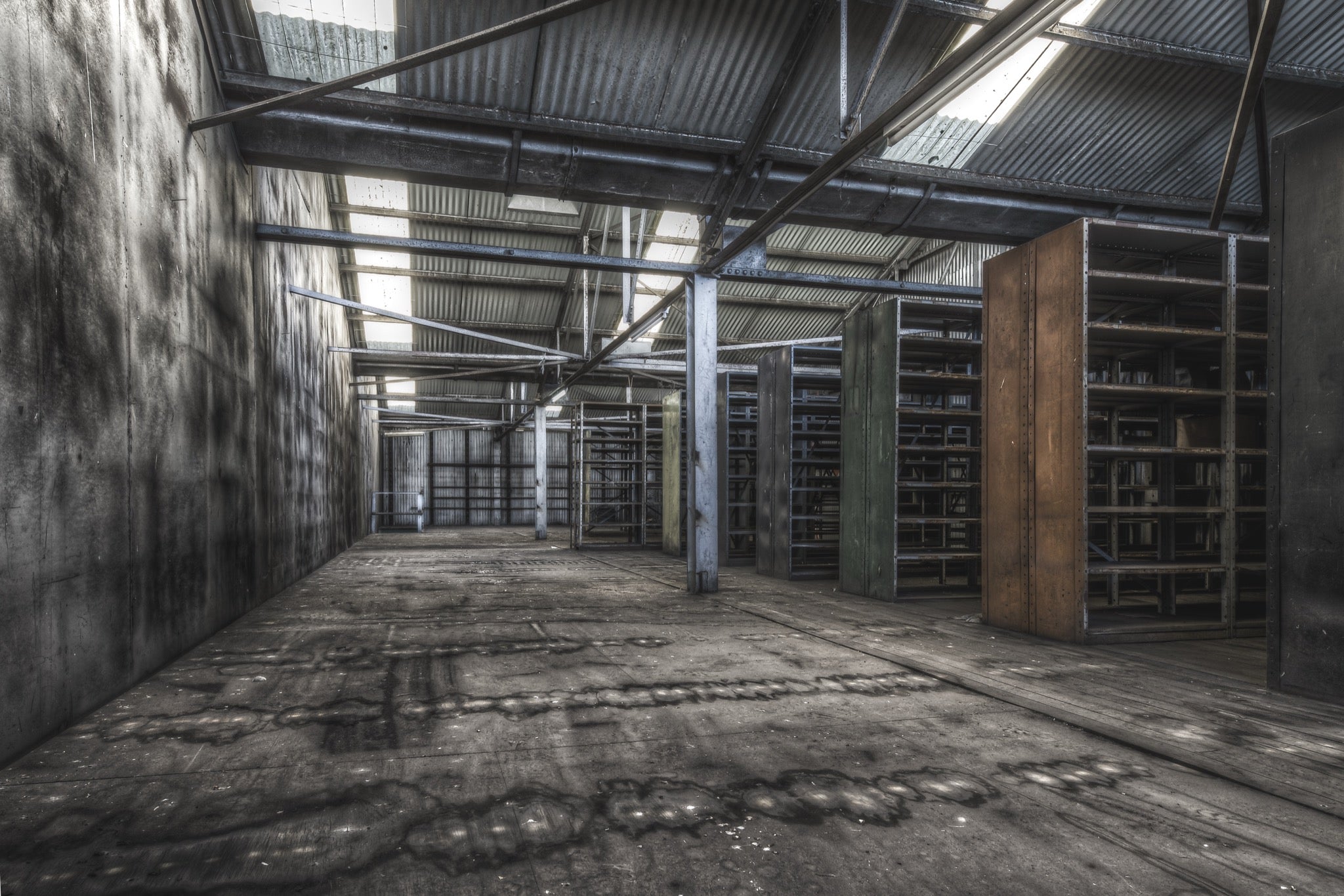 Emptied metal shelving units receding through the upper storeroom at Awaba Colliery, with skylights in a corrugated iron roof casting light across water-stained concrete walls and worn timber floorboards.