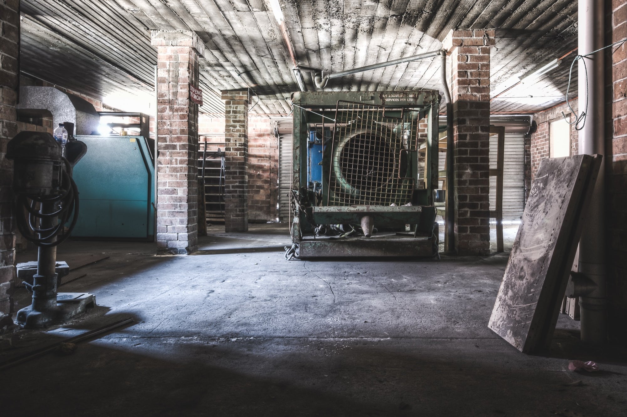 A large vent fan in green mesh caging in a basement workshop at Awaba Colliery, with a partial red danger sign on a brick column and a drill press with coiled hoses in the foreground.