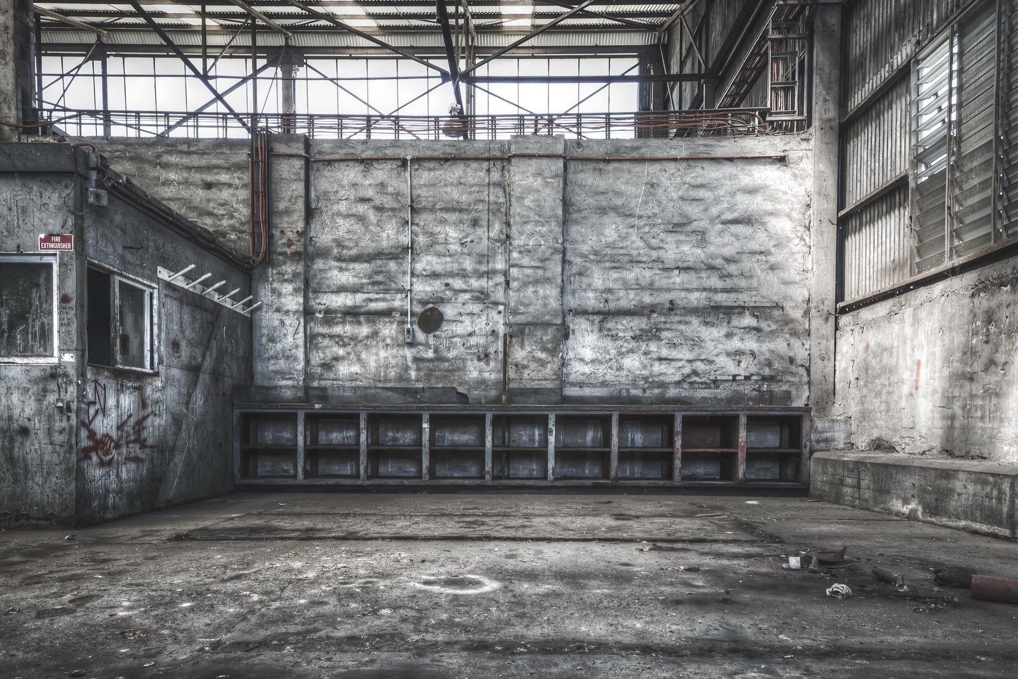 Empty shelving units and a faded fire extinguisher sign on the rendered wall of a workshop bay at Awaba Colliery, with graffiti near two windows and exposed pipes overhead.