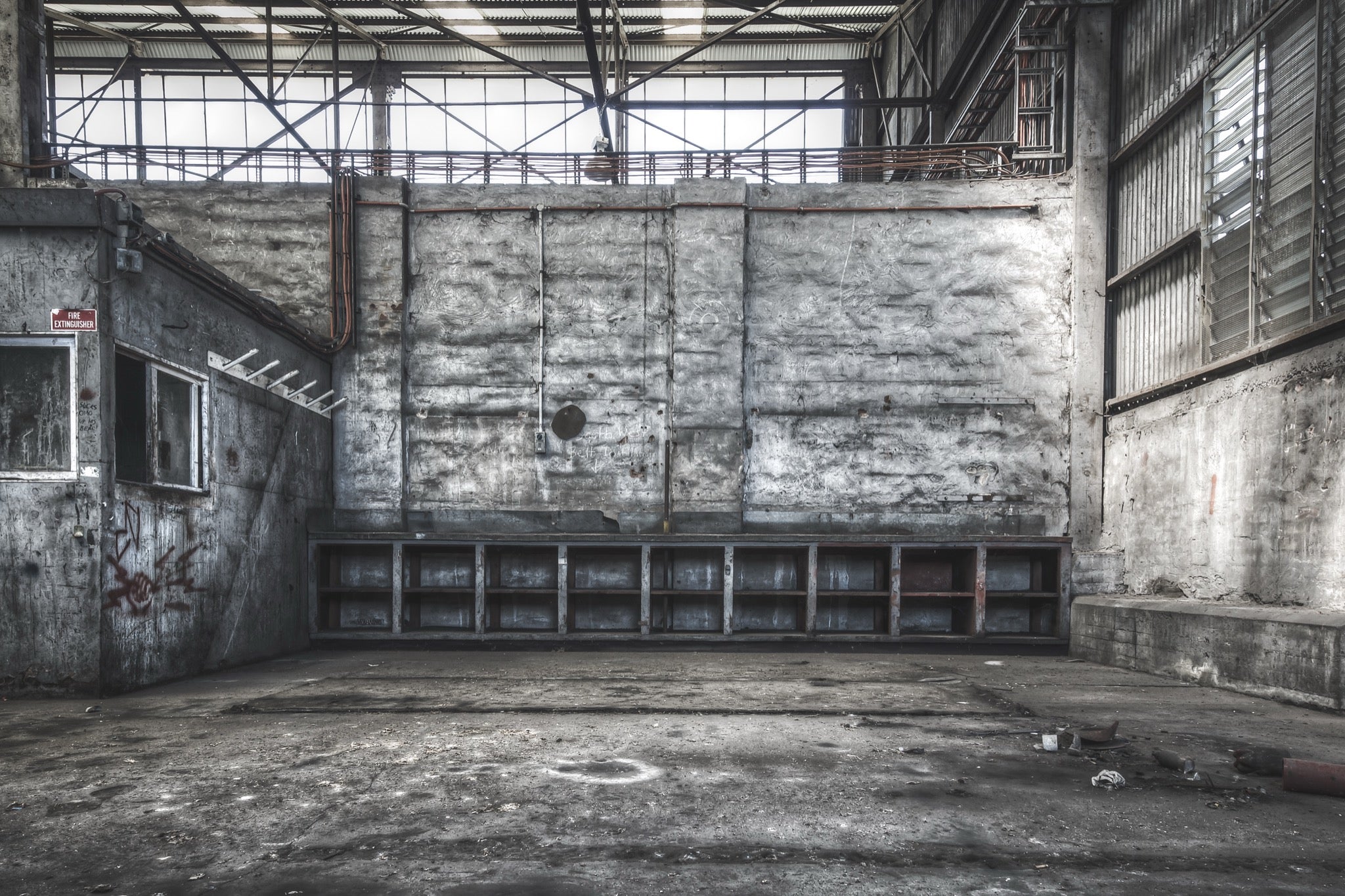 Empty shelving units and a faded fire extinguisher sign on the rendered wall of a workshop bay at Awaba Colliery, with graffiti near two windows and exposed pipes overhead.