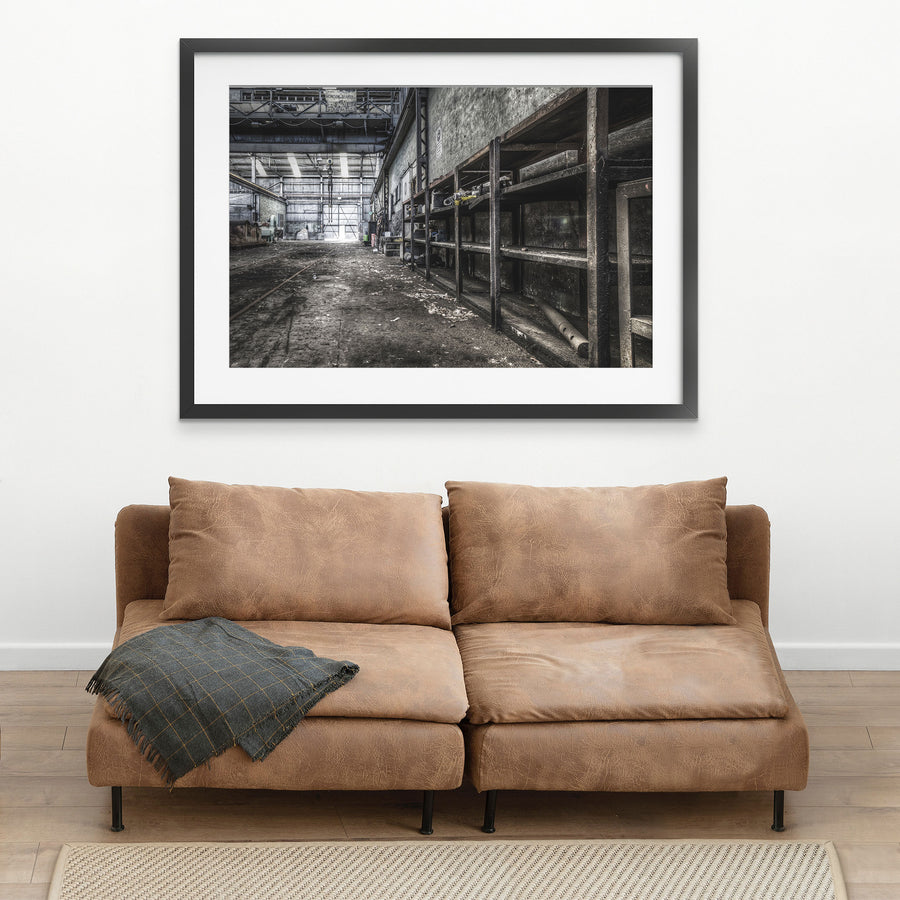 Rusted workshop shelving and faint floor rail tracks inside the colliery workshop at Awaba, with a crane bearing the name "Morison Bearby" visible overhead and a white cup resting on the concrete floor.