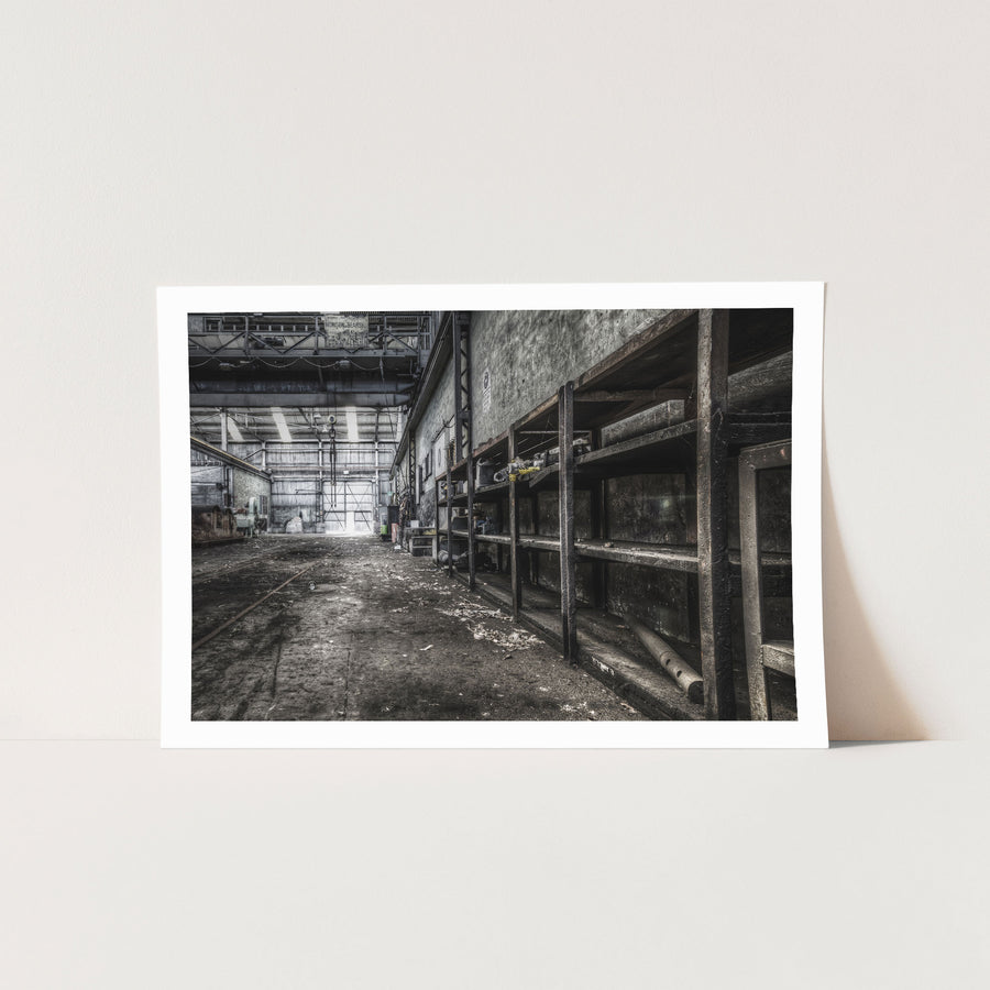 Rusted workshop shelving and faint floor rail tracks inside the colliery workshop at Awaba, with a crane bearing the name "Morison Bearby" visible overhead and a white cup resting on the concrete floor.