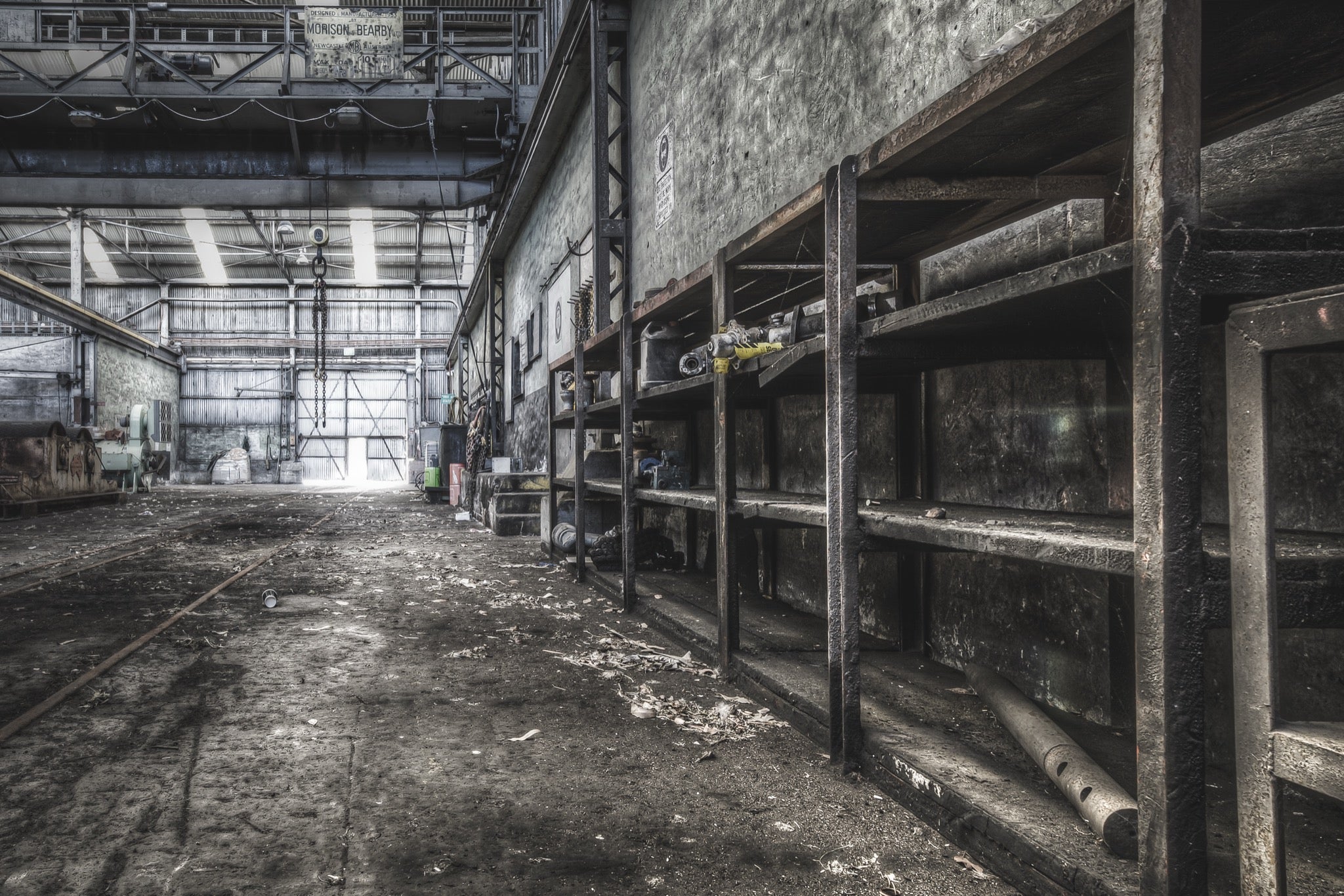 Rusted workshop shelving and faint floor rail tracks inside the colliery workshop at Awaba, with a crane bearing the name "Morison Bearby" visible overhead and a white cup resting on the concrete floor.