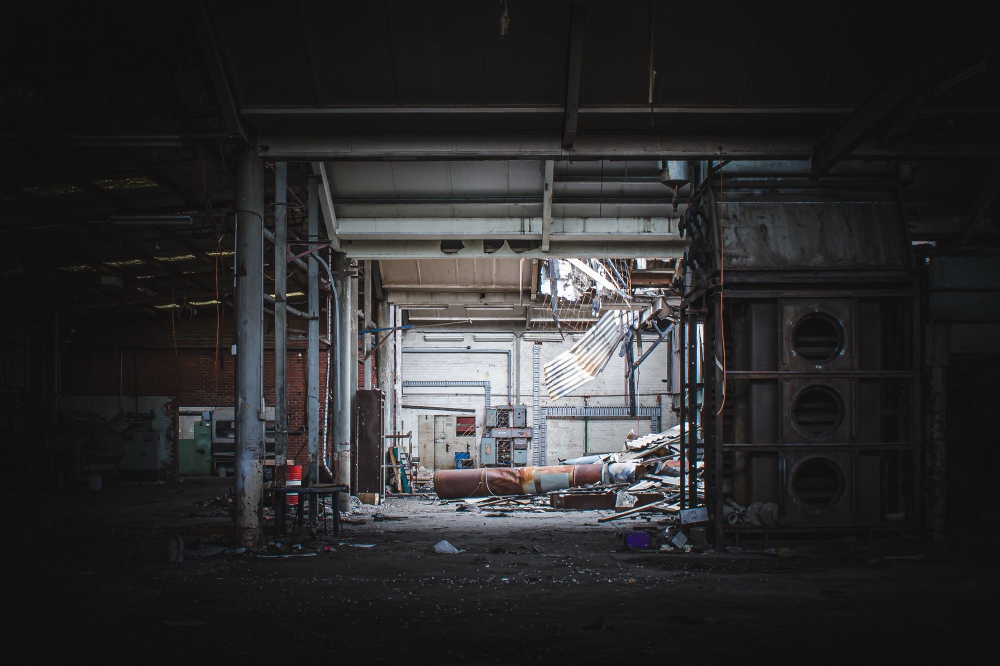 A collapsed sheet-metal roof section hanging across the rear wall of a derelict manufacturing hall at the former Davies Coop / Bradmill factory in Yarraville, with a large rusted cylindrical pipe and scattered debris on the concrete floor.