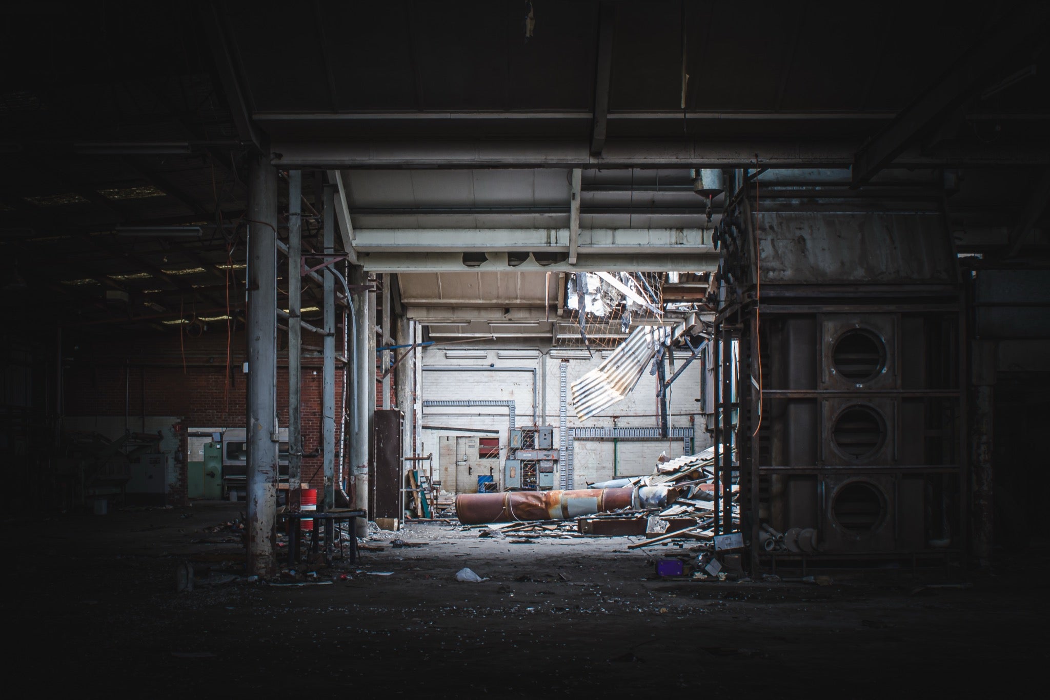 A collapsed sheet-metal roof section hanging across the rear wall of a derelict manufacturing hall at the former Davies Coop / Bradmill factory in Yarraville, with a large rusted cylindrical pipe and scattered debris on the concrete floor.